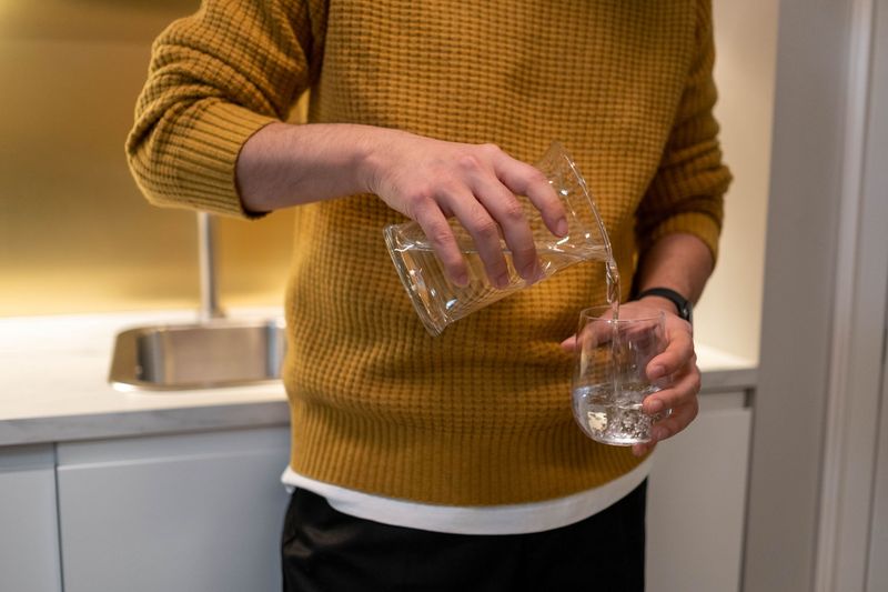 Person i gul genser heller vann fra en mugge i et glass på kjøkkenbenken