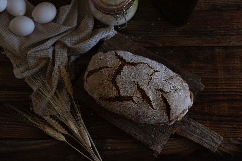 Rundt rugbrød med sprukket skorpe på trebrett, ved siden av hveteaks