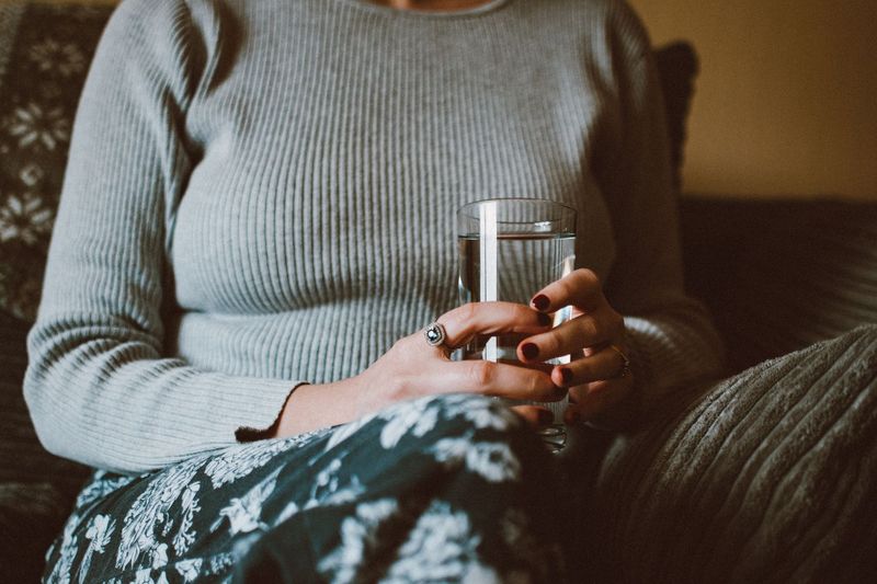 Kvinne i strikkegenser holder et glass vann i fanget på en sofa
